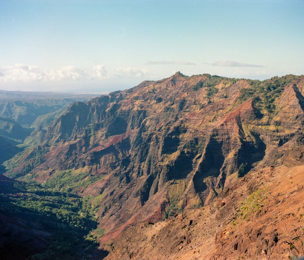 A photograph of Waimea Canyon taken on a Pentax 6x7 and Kodak Portra 160 film stock. In this photo, a massive mountain can be seen rising above the green riverbed at the bottom of the canyon. 