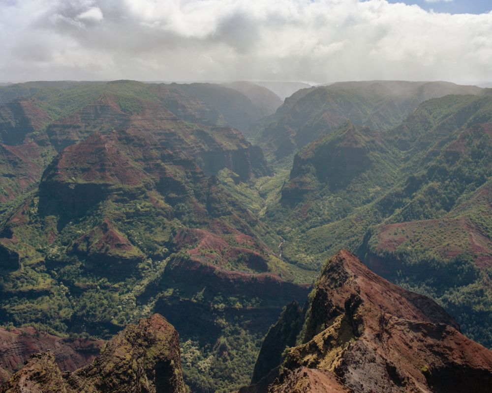 A photograph of Waimea Canyon taken on a Pentax 6x7 and Kodak Portra 160 film stock. In this photo, the tall canyon walls are seen towering above the rivers running wildly through the bottom of the canyon. 