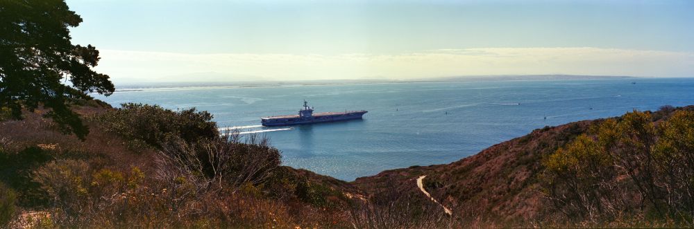 A 6x17 panorama of a US Navy Ship leaving North Island headed into the Pacific Ocean. The ship tears through the water, framed by the tall hills of Point Loma. 