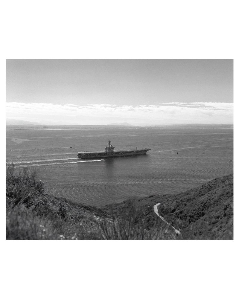 A black and white photograph of a US Navy ship leaving North Island en route into the Pacific Ocean. On either side of the image, the cliffs of Point Loma are seen high above the water. 