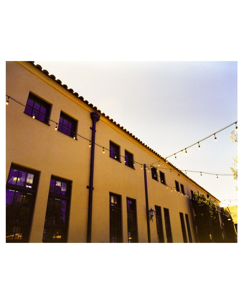 A color photograph of a Spanish-style building at the Liberty Public Market in San Diego, CA. The adobe building dominates the frame with outdoor pendant lights strung throughout the image. 