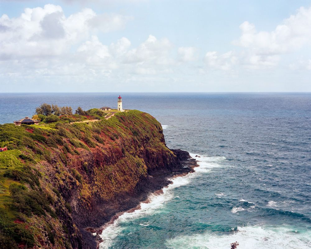 A photograph of the Kilauea Lighthouse located on the island of Kaua'i. In this photo, the lighthouse is seen on the edge of a rocky outcropping standing high above the waves crashing into the rocks below. 