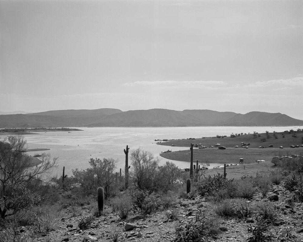 A black and white photograph of Lake Pleasant, taken just outside of Phoenix, AZ. In the foreground, common desert plants can be seen (like the saguaro) while the lake dominates the rest of the image. In the background, mountains are seen rising over the lake. 