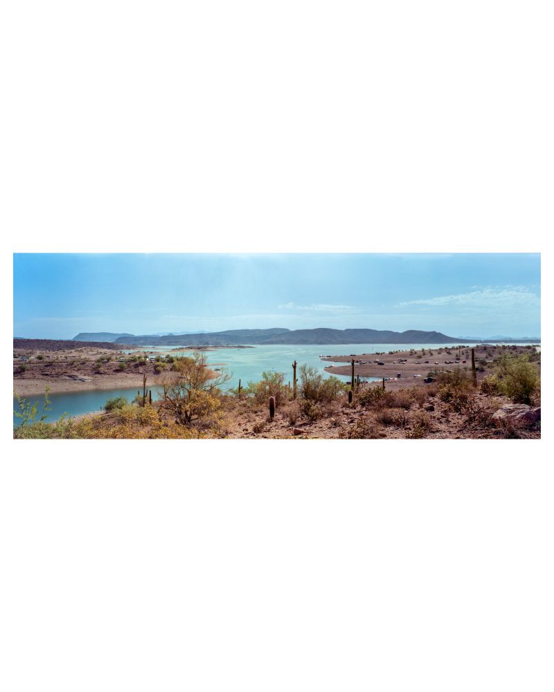 A panoramic photo of Lake Pleasant just outside of Phoenix, AZ. In the photo, cars are seen parked on the banks of the lake as locals recreate on the shores. 