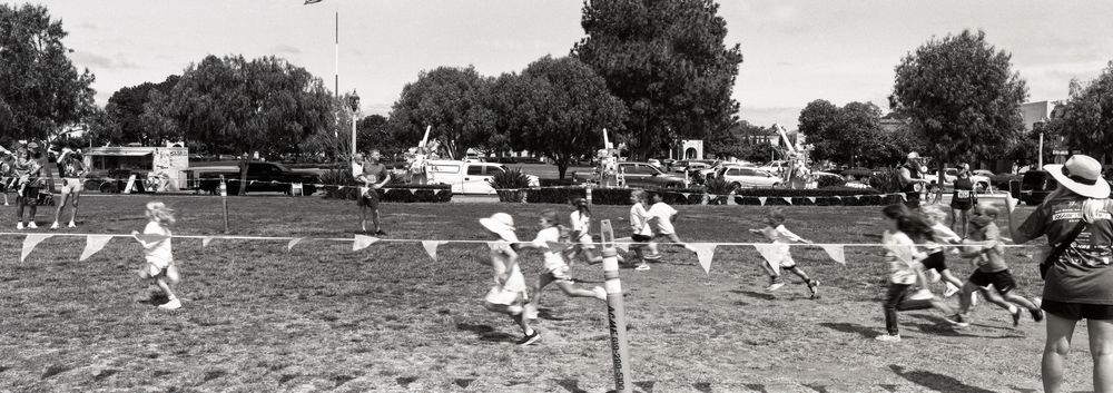 A black and white panoramic image of children competing in a race. The children are blurred as they run past the camera. 