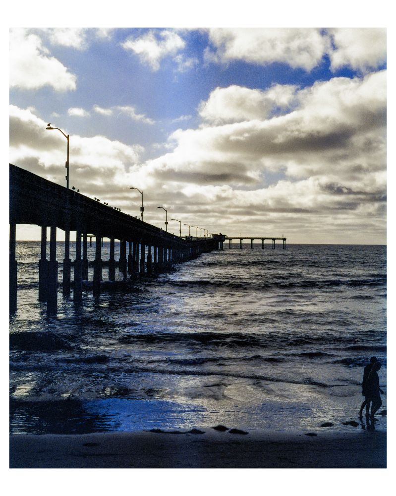 A color photograph of Ocean Beach Pier in San Diego, CA. A shadowy pier stretches into the sunset as a man and woman walk out of frame along the beach. 
