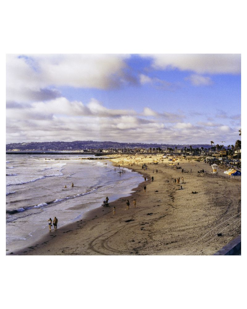 A color photograph of Ocean Beach in San Diego, CA. Locals and tourists alike flock to the beach to enjoy the sunset as waves crash onto the shore. 