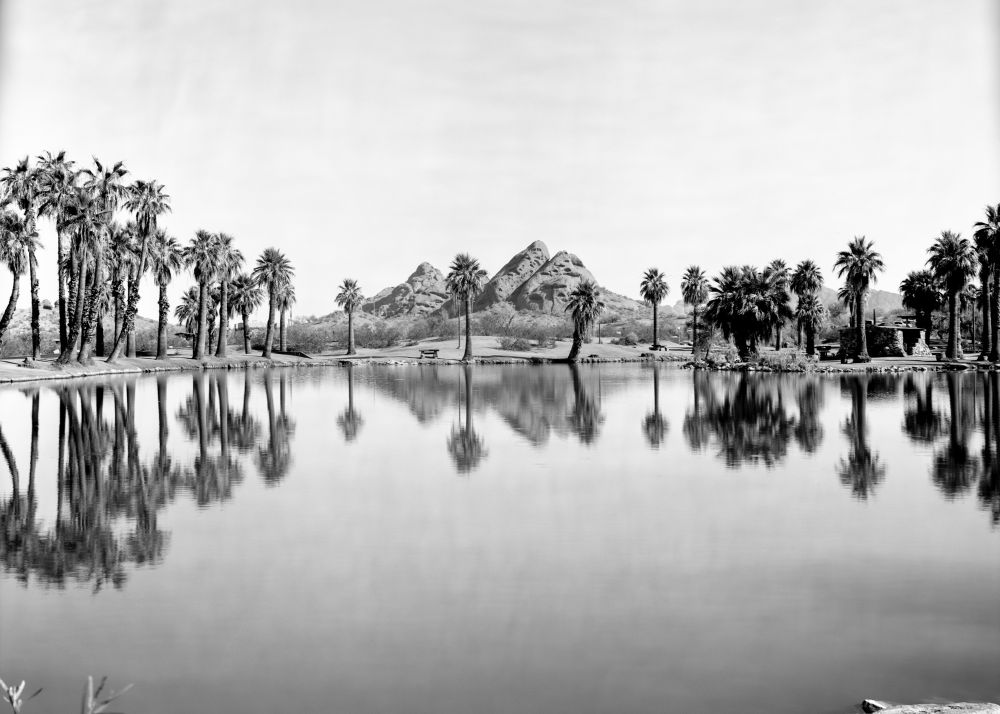 A black and white photograph taken at Papago Park in Phoenix, AZ. The red rock formations are central to the image. In the foreground,  fishing pond can been seen lined with palm trees that reflect in the water. 