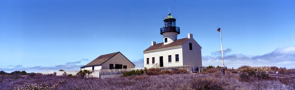 A 6x17 panorama of the Old Point Loma Lighthouse. In the image, the lighthouse and auxiliary building are seen built from a white-painted brick. An American flag flies next to the buildings. 