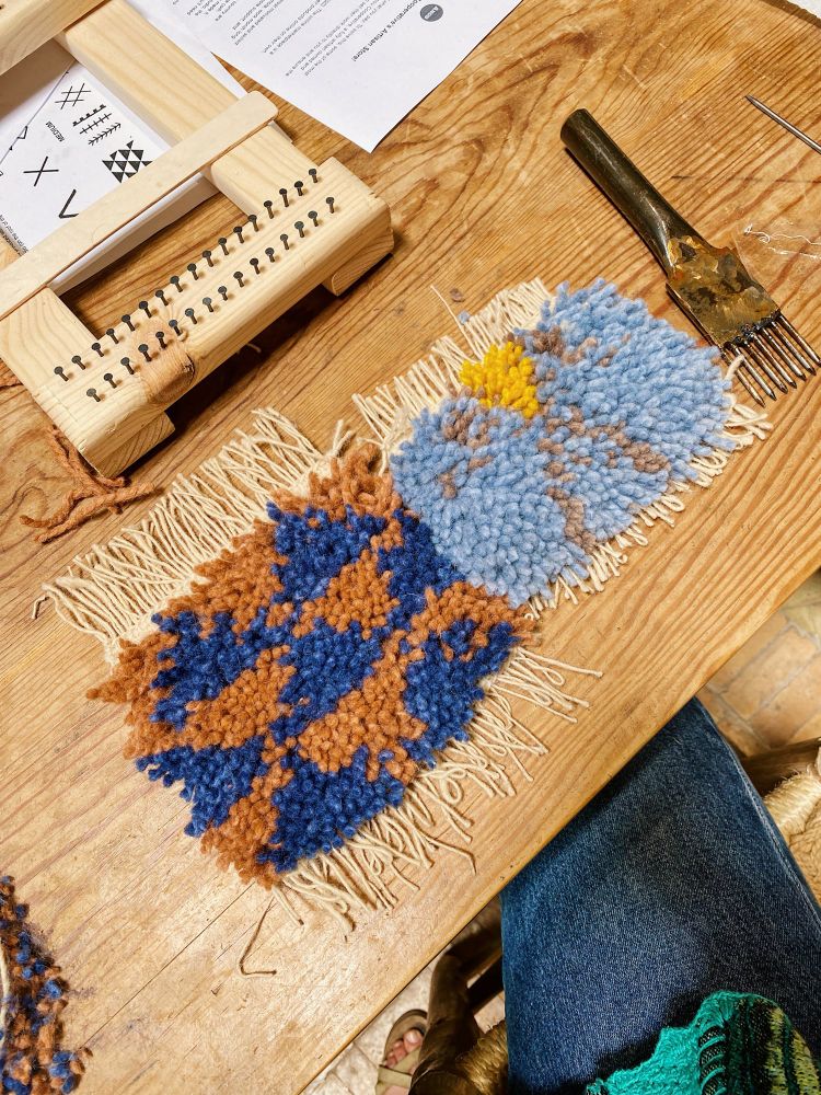 My finished mini rug next to Ben’s, alongside one wooden loom and a metal weaving comb. The design of mine is blue and terracotta triangles. Ben’s rug has a light blue background with violet diagonal lines forming a central diamond, which is bright yellow.