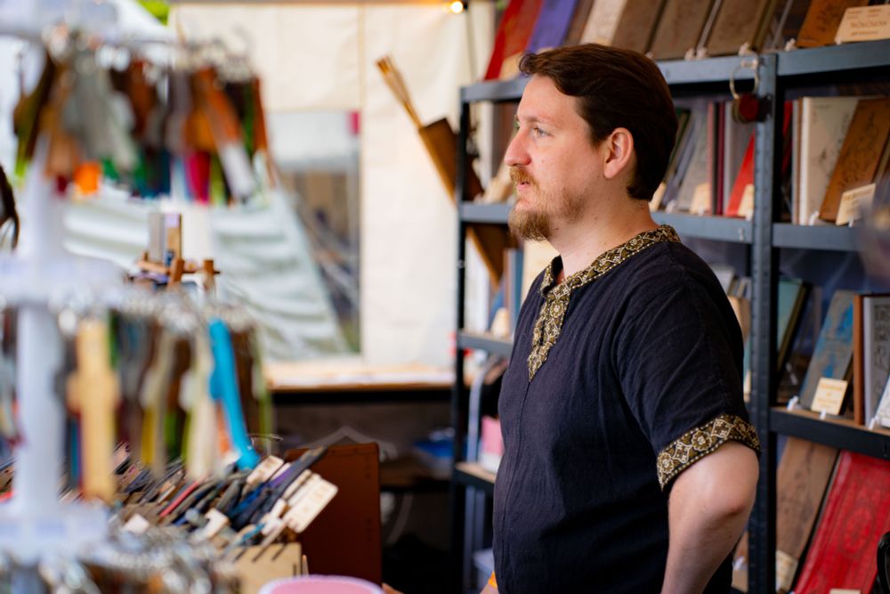 A man in black tunic standing in his tent at a convention, with a wall of leather notebooks behind him.