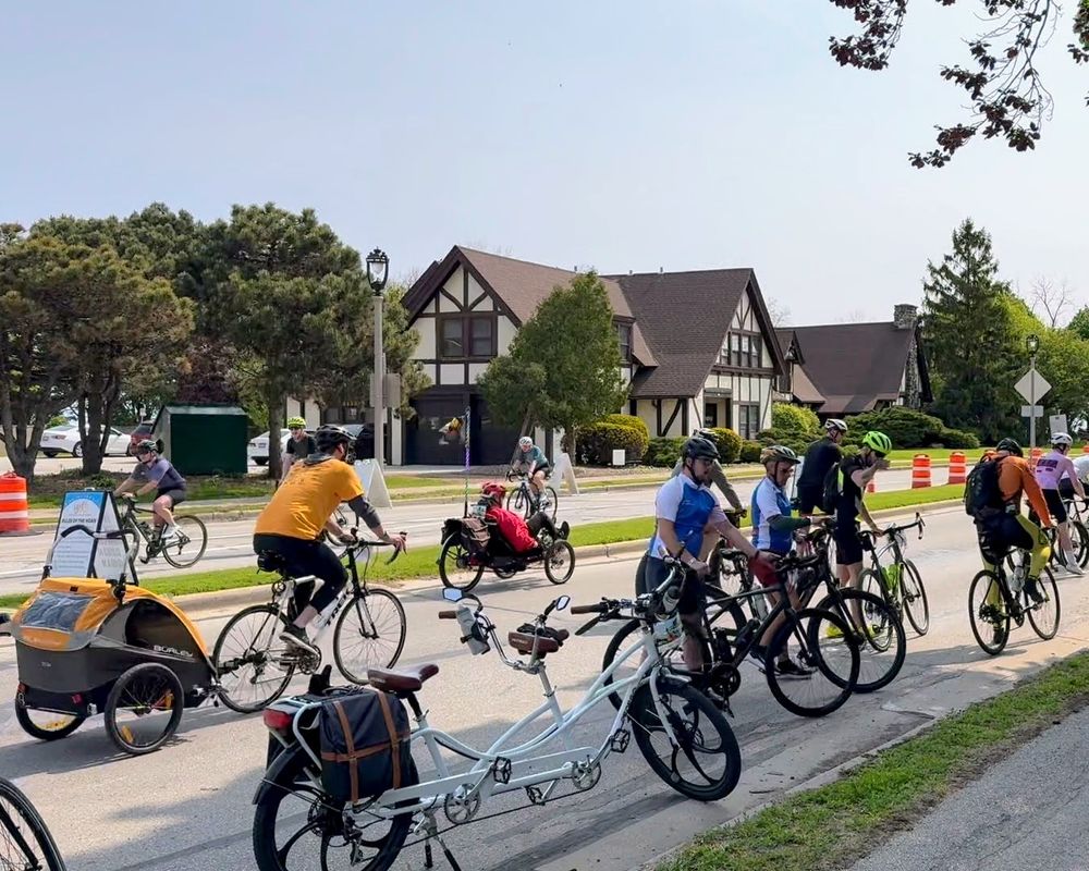 a rest stop on the bike ride with many types of bikes visible including a trailer, tandem, and recumbent. A tudor style park fieldhouse is in the background