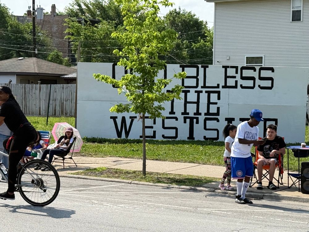 Parade goers watching from the sidewalk in folding chairs with the mural in the background that says “god bless the westside”