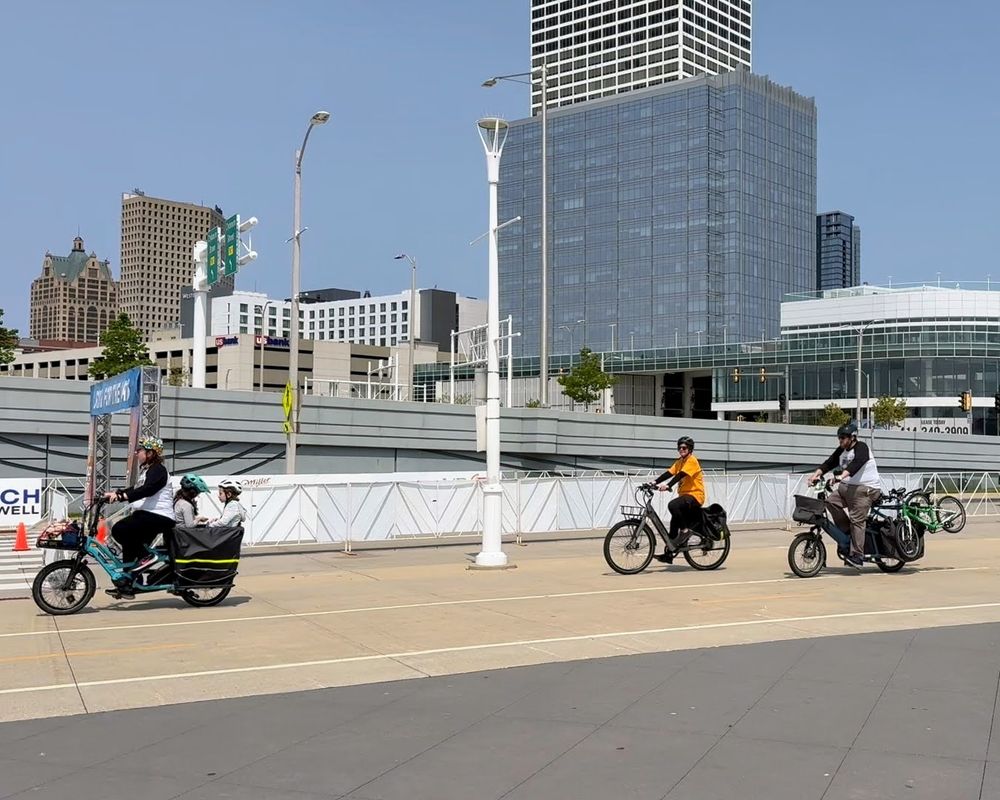 People riding on the Milwaukee lakeshore path with the downtown skyline in the background. The people are riding cargo bikes, one bike has two kids in tow and the other cargo bike has two kids bikes attached in the cargo section