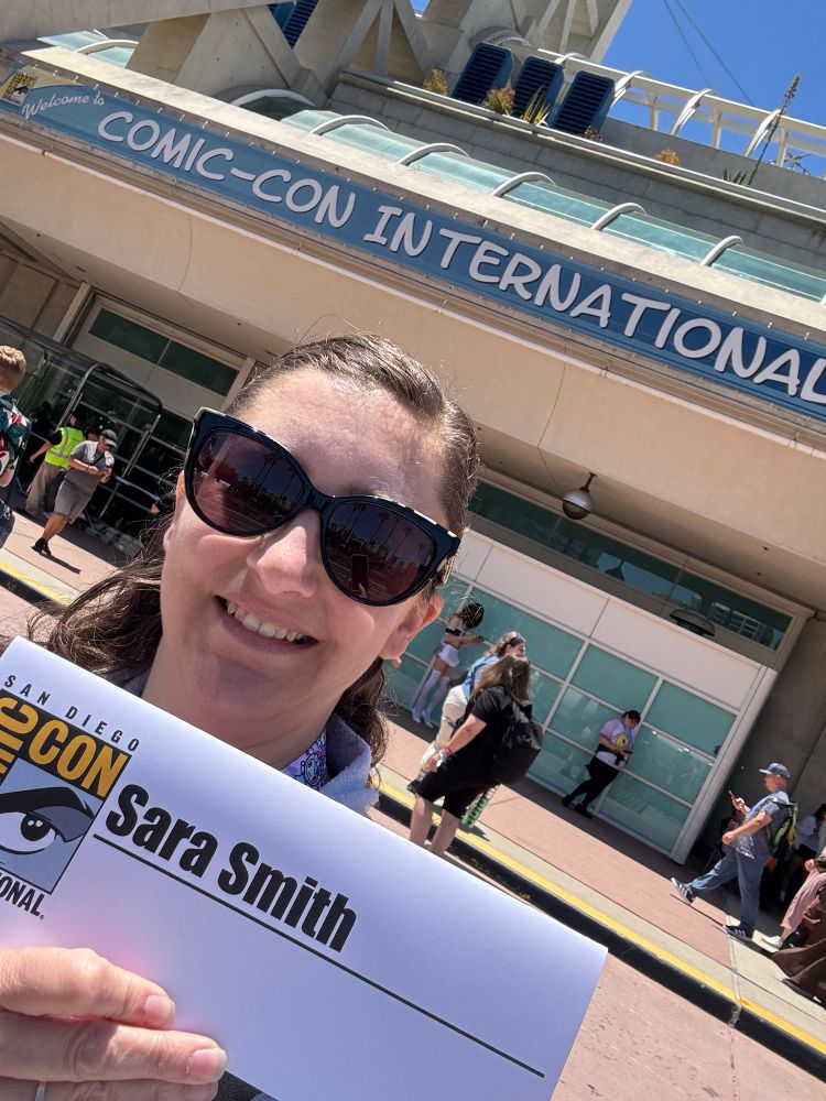 A woman holds her comic con name plate reading “Sara Smith” while smiling and standing in front of a sign that reads “Comic Con International”