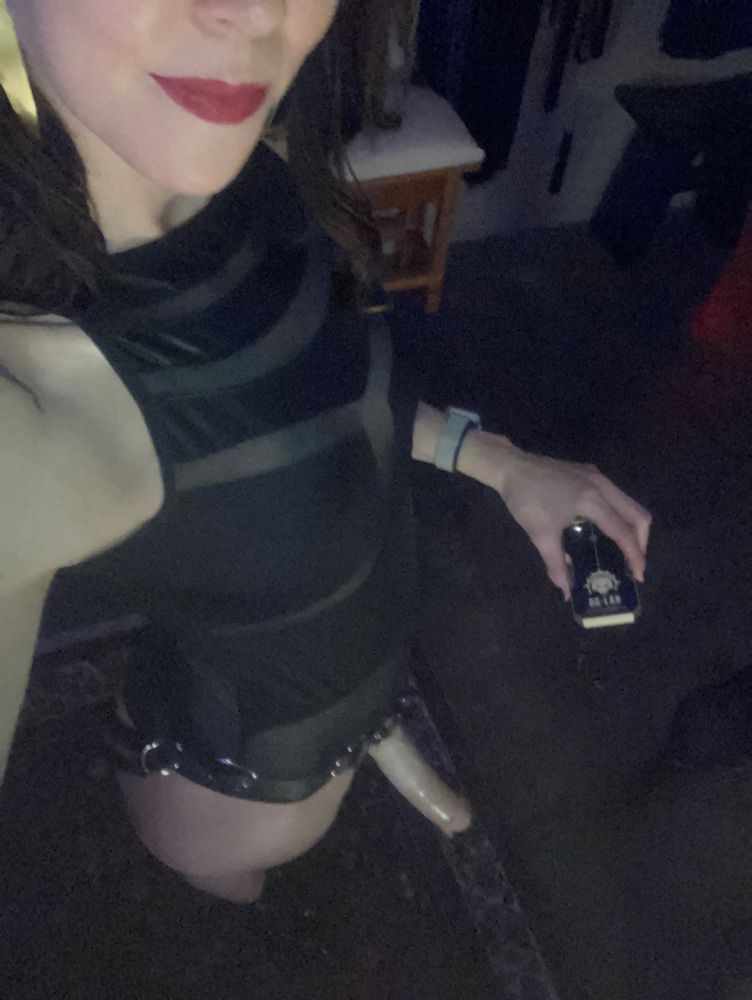 Mistress Mercury is seen taking a selfie from the nose & mouth down. She is tall with pale skin, a sexy black bodysuit, a black rubber strap on harness with a large cock in it, and a knowing grin on her face as her other hand holds an electro stim box.