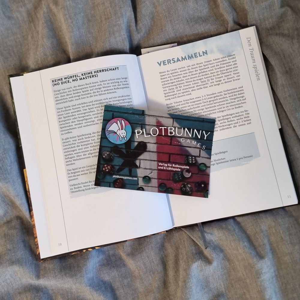 A book page from Dream Askew in German and a leaflet from Plotbunny Games on top of it. 