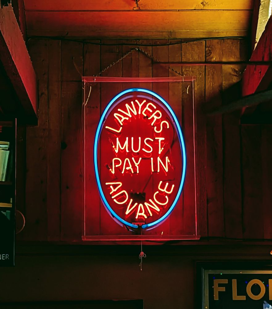 A bar sign hanging on a wooden wall, spelling out brightly in neon, "Lawyers must pay in advance"