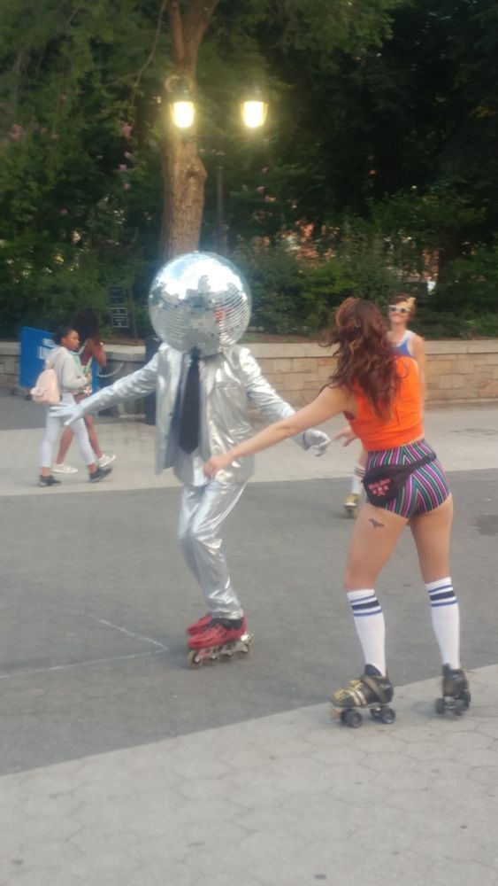 A wide concrete area on the edge of a park, with people walking around and two people on roller skates: A woman in an orange blouse and snug striped shorts, and a man in a shiny silver suit and black tie whose head is inside a mirrored disco ball