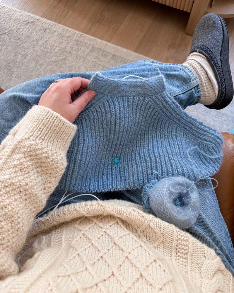 Woman sitting cross legged in cream cable knit sweater and blue denim with blue handknit sweater in progress in lap.