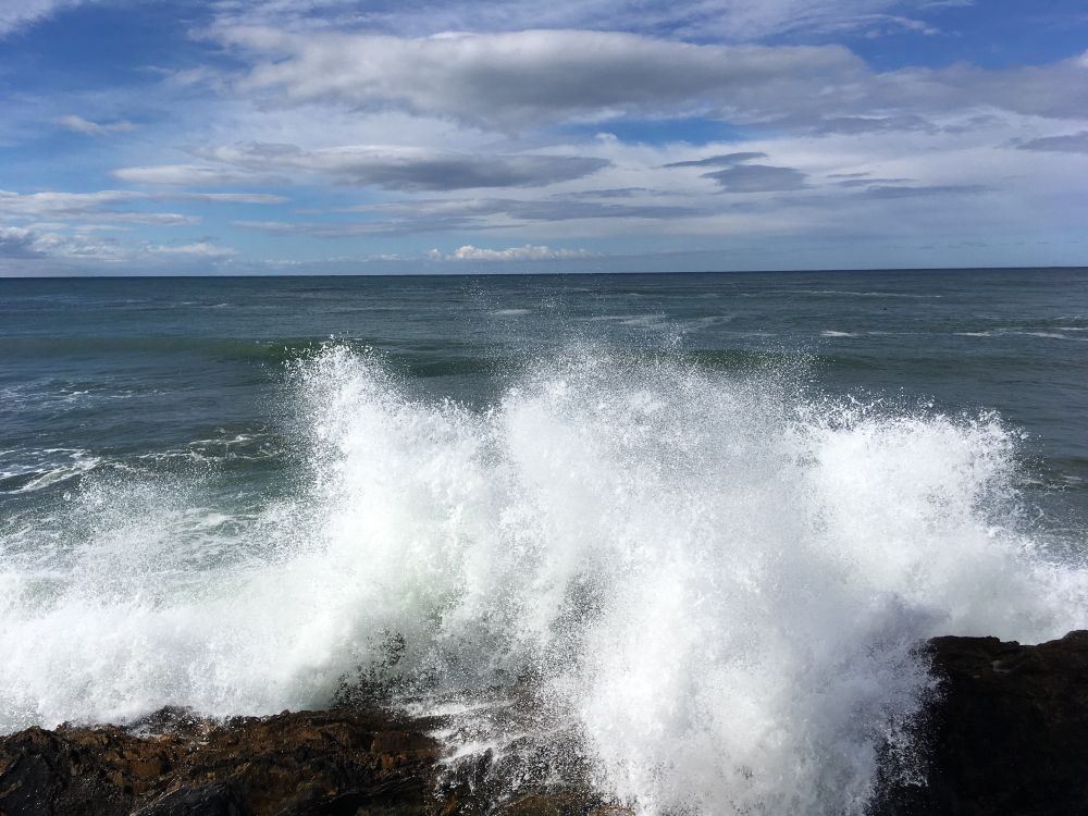 Last moment the wave’s splash begins to flow over wet rocks the sea is a dark teal color in the morning light that filters through clouds in a blue sky.