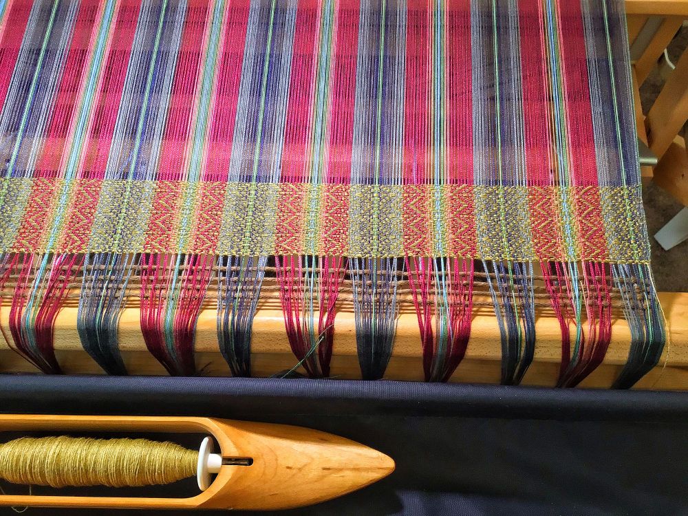 The colors are reds grays and blue teen woven with a golden weft. On the loom.