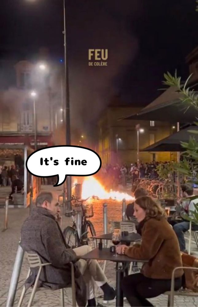 Photo of 2 people chatting in a Paris cafe while a fire burns in the middle of the street from protestors
