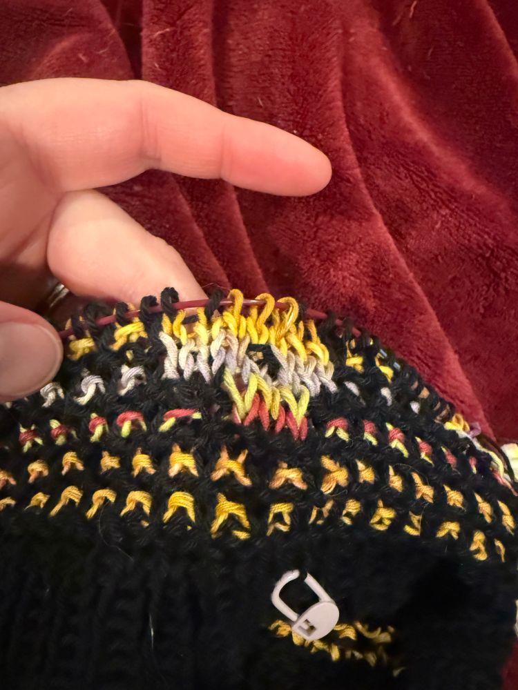 The start of a new sweater is held by Annie’s fingers. It is a black mosaic knitted yoke with a yellow, purple, red, green, and white yarn forming the dots and flowers portion of the pattern. 