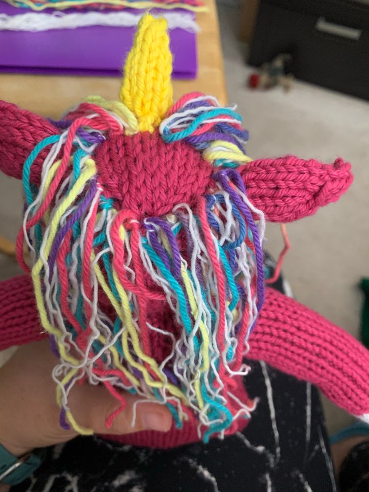 The top of a head of a pink knitted unicorn, in the midst of knotting on the mane. The mane is multicolored and white yarn. 