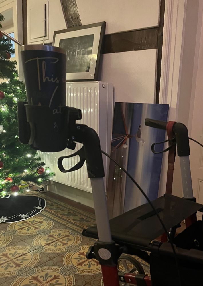 Foto meines Rollators, der jetzt einen Kaffeebecherhalter am Griff hat. Im Hintergrund sind eine Heizung, der Teil eines Weihnachtsbaums, sowie zwei Bilder (eines mit einer Möwe auf einem Ruderboot am Meer (an der Wand lehnend) und eine schwarze Fotografie ( auf einer Heizung stehend) einer Bekannten von mir. Aufgenommen im Flur eines alten Bauernhauses mit Fachwerk