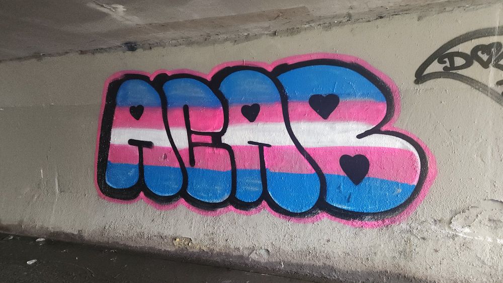 ACAB in the blue, pink, and white of the trans flag in big bubble letters with heart counters painted on a pedestrian underpass.
