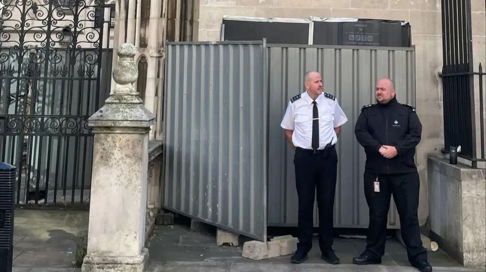 Two uniformed security officers stand in front of a grey corrugated metal barrier hastily erected against a stone wall, blocking the view of where the street art had been. The scene suggests an official attempt to conceal or suppress the mural.
