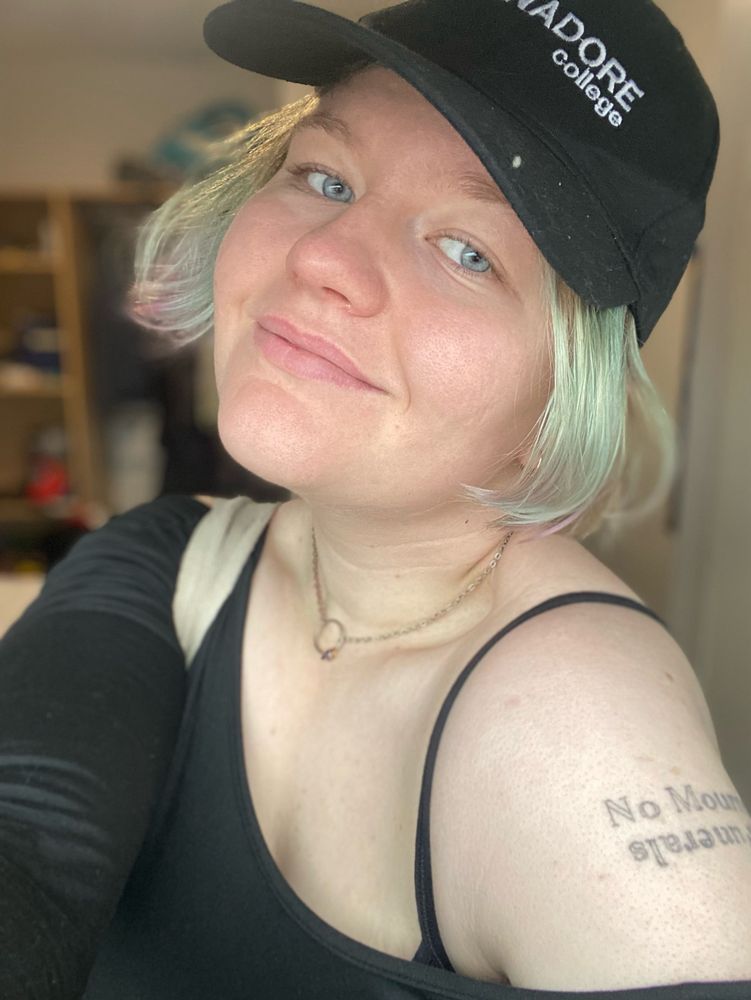 A selfie of a person with chin length blue hair. They have a tattoo on their shoulder and are wearing a shoulderless top and a black ball-cap 