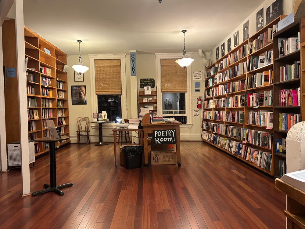 The Poetry room of City Lights Booksellers. Full bookshelf’s on both sides of room. Table in middle with books stacked on top. Sign that says Poetry Room!