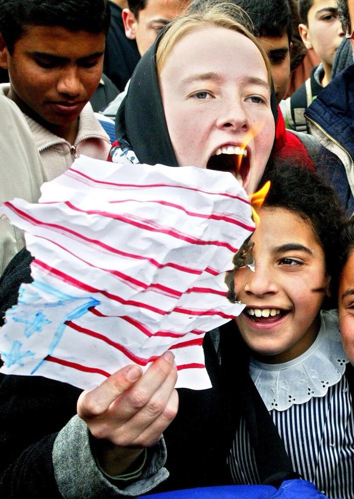 rachel corrie, a young white american woman surrounded by palestinian children and young people, holding an american flag drawn on paper held upside down and set on fire. she was murdered by the israeli army at 23.  