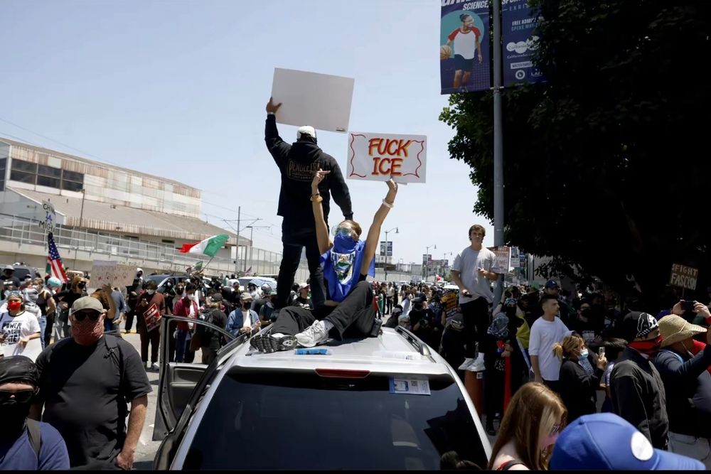 crowd of protesters in LA with two sitting in the top of a parked car holding signs that say FUCK ICE