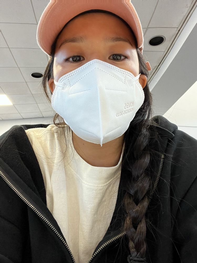 woman with pink baseball cap, long dark side braid, and and white mask seated wearing a white t shirt and black hoodie
