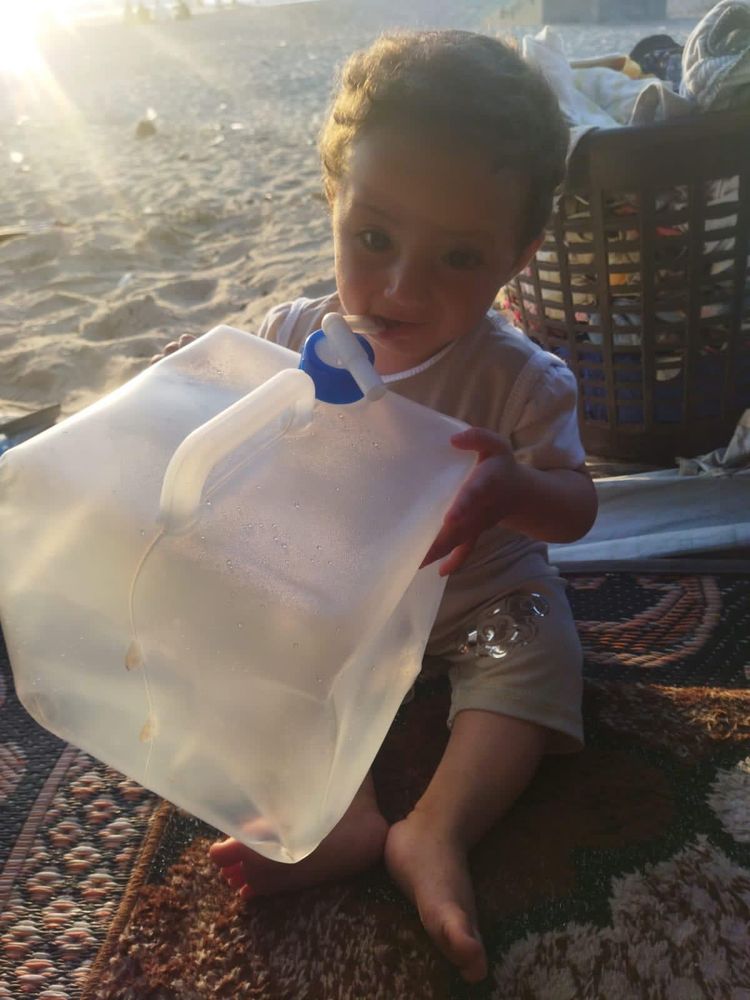 baby hamoud, a toddler with curly brown hair and big brown eyes wearing a t shirt and shirts holding an empty plastic water jug while sitting on a carpet in the sand 