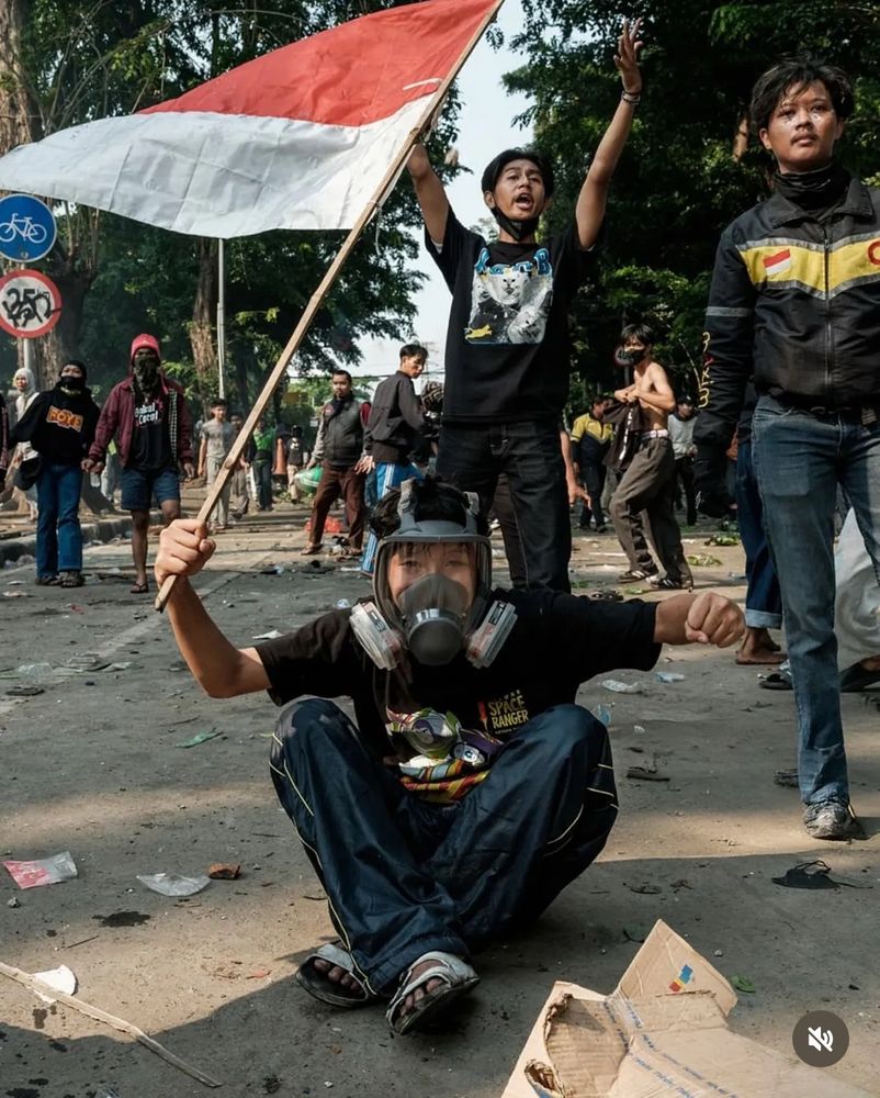 a young protester sits on the ground wearing a gas mask and waving an indonesian flag 