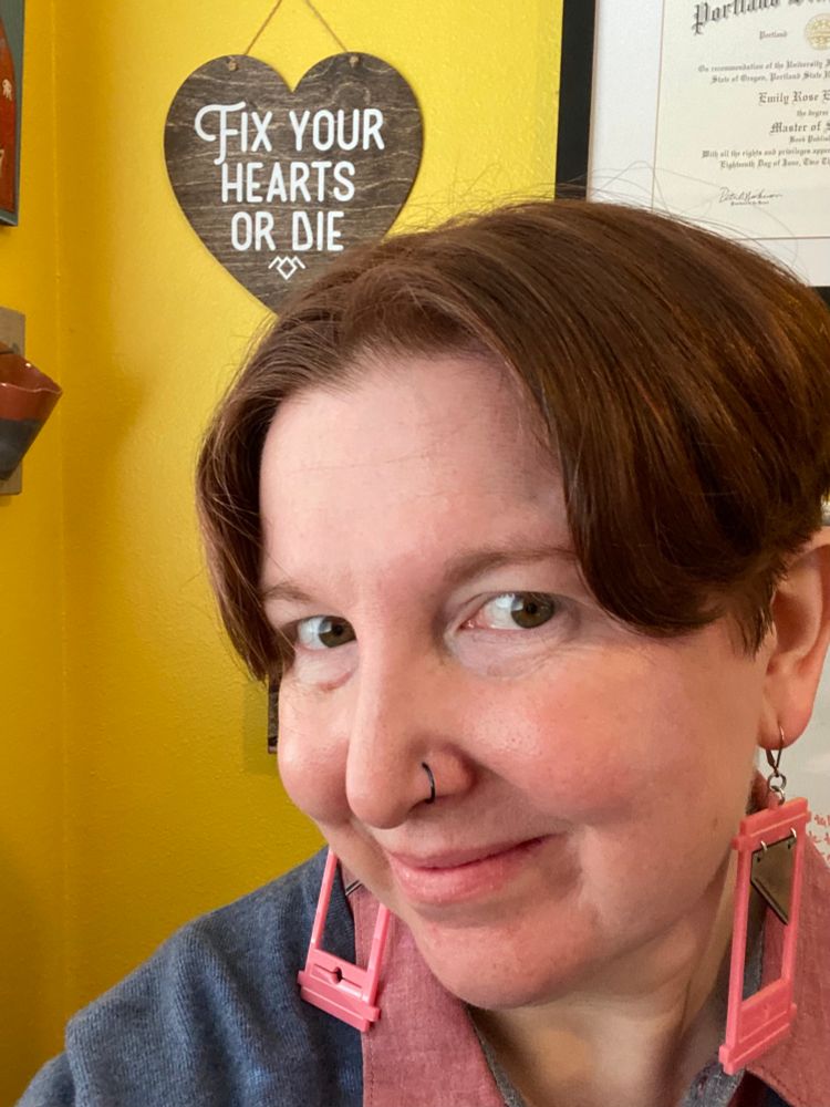 Person with short red hair parted in the middle, pink guillotine earrings, and a blue sweater, smiling in front of a heart shaped wall sign that says “fix your hearts or die”