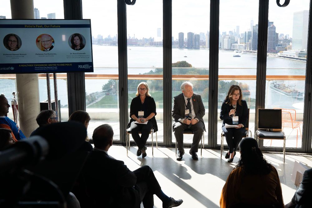 Panel discussion at an event with three speakers seated in front of a large window overlooking a city skyline, with attendees listening attentively. 