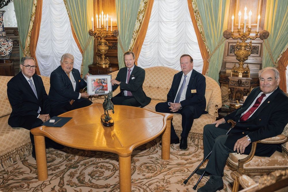 Five individuals seated around a table in an ornate room, holding a framed artwork.