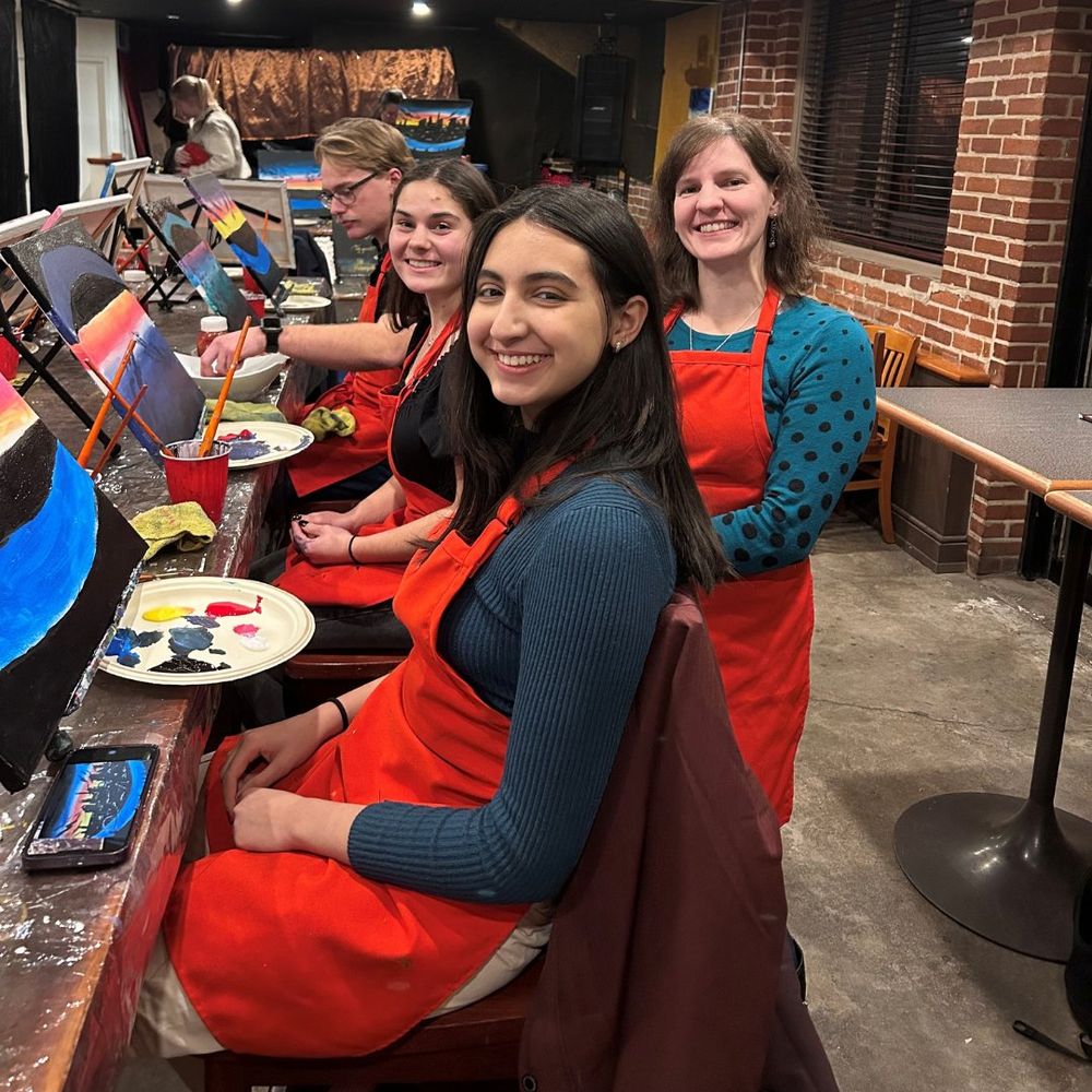 Four people wearing aprons sit at a table with painting supplies, smiling at the camera in a casual art class setting.