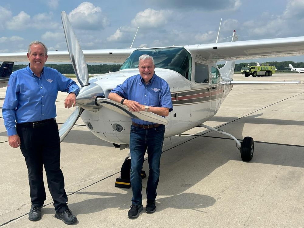Two individuals in blue shirts smiling next to a Cessna aircraft on a sunny tarmac.