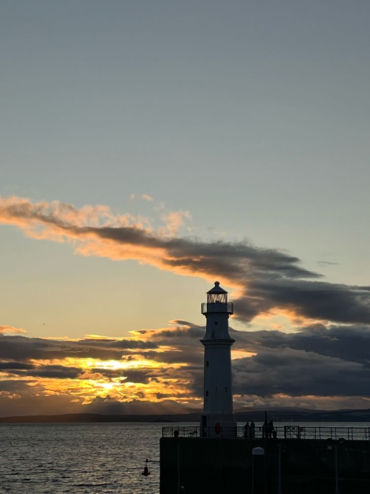 The harbour at Newhaven in Edinburgh at sunset with shreds of clouds aglow in the dying sunlight and the lighthouse a near silhouette. 