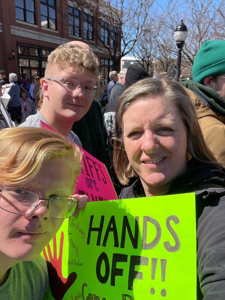 Janet, a white woman holds up a Hands Off sign and is surrounded by her white sons and other protesters.