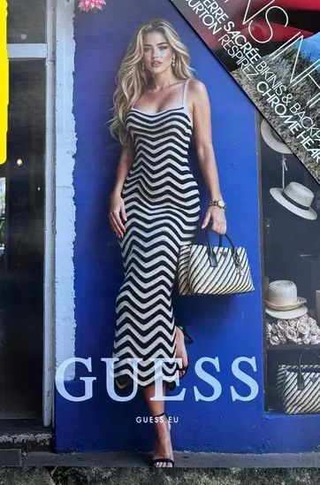 An AI generated image of a blond woman wearing a black and white horizontal striped dress in front of a blue wall. She appears to have legs the length of which would look more appropriate on a literal Barbie doll.