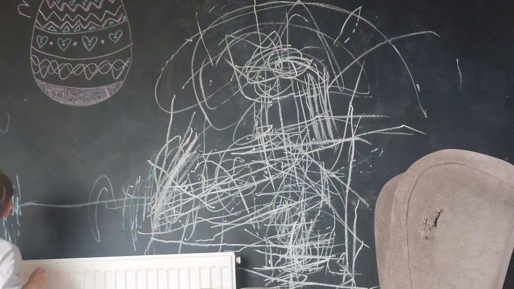 A chalkboard wall covered in white and blue scribbles. The ear and shoulder of said hyperactive toddler is visible in the bottom right corner