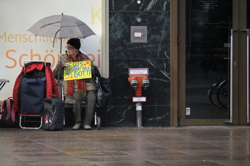 En äldre kvinna sitter på en stol. Hon har ett paraply över huvudet och en skylt där det står ”Jesus versöhnt dich mit Gott!“