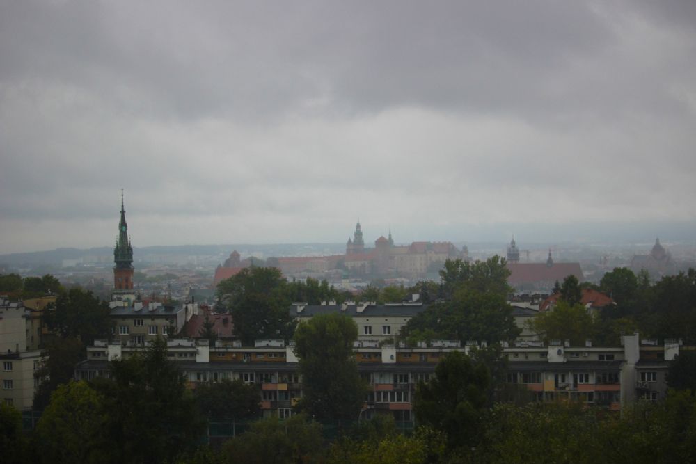 Ett grått och regnigt Krakow. Gråa moln på himmeln, takåsar och kyrktorn  och trädtoppar i förgrunden. 